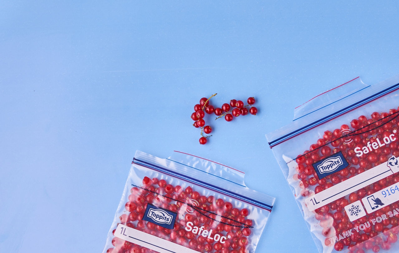 Zwei Toppits SafeLoc Gefrierbeutel mit eingefrorenen roten Johannisbeeren liegen auf einer blauen Fläche, daneben einige frische Johannisbeeren 